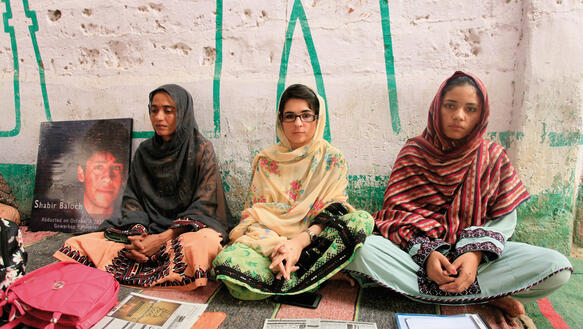 Drei junge Frauen in Gewändern und Kopftüchern sitzen im Schneidersitz auf dem Boden und blicken in die Kamera