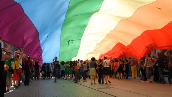 Viele Menschen demonstrieren auf einer Straße unter einer riesigen Regenbogen-Fahne