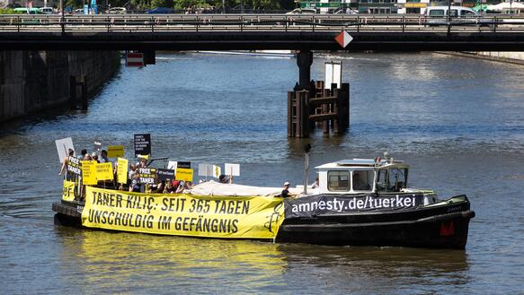 Ein Boot auf einem Fluss mit mehreren Menschen die Schilder hochhalten, an der Seite hängt ein Banner auf dem steht: "Taner Kılıç seit 365 Tagen unschuldig in Haft"  