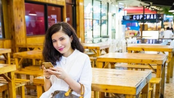 Frau sitzt mit ihrem Smarthphone auf einer Bank und lächelt in die Kamera