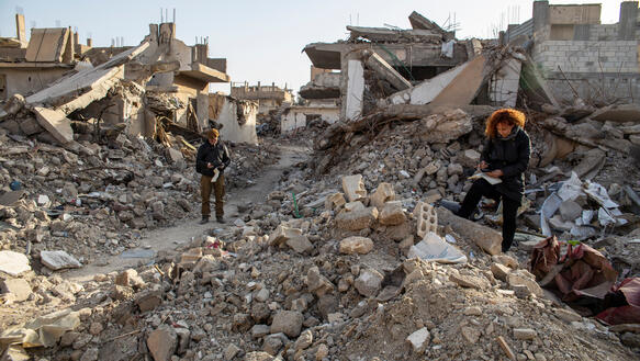 Ein Mann und eine Frau stehen inmitten von Trümmern und Ruinen zerstörter Häuser und machen sich Notizen