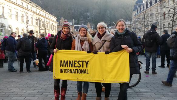 Vier junge Frauen in Winterkleidung stehen in einer gut gefüllten Fußgängerzone und halten ein gelbes Banner vor sich, auf dem "Amnesty International" steht, im Hintergrund s