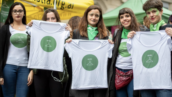 Fünf junge Frauen halten weiße T-Shirts hoch auf denen ein grüner Kreis mit einem weißen Kleiderhaken zu sehen ist 