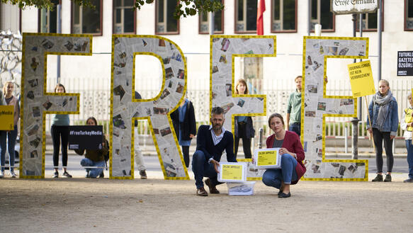 Mehrere Personen halten vier meterhohe Buchstaben, die das Wort "Free" bilden, davor hocken zwei Personen mit Boxen voller Petitionen 