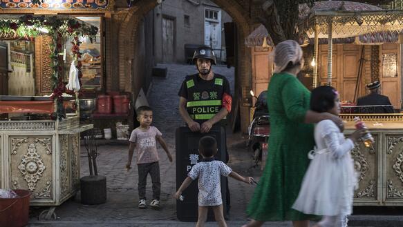 Ein Polizist mit Schild steht vor einem Tordurchgang, um ihn herum zwei Kinder, davor eine Passantin mit Kind