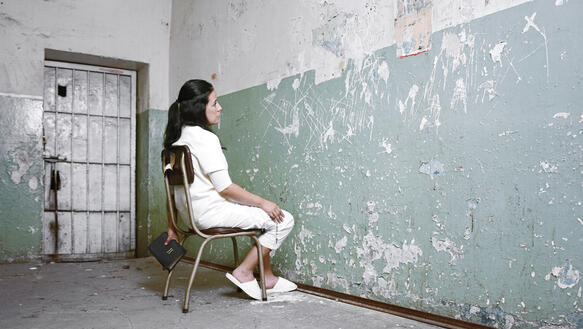 Eine Frau sitzt in einer Zelle auf einem Stuhl und starrt die Wand an