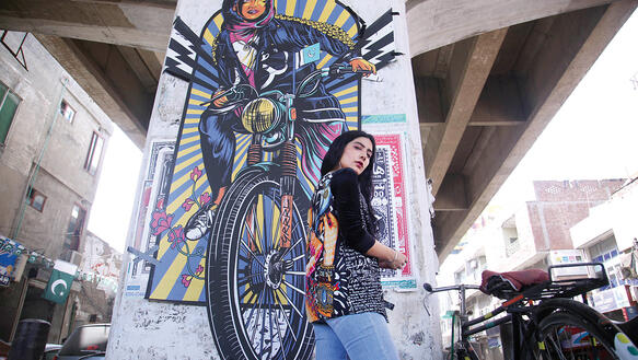 Eine Frau mit Jeans und langem schwarzem Haar steht vor einem Graffiti einer Frau auf einem Motorrad