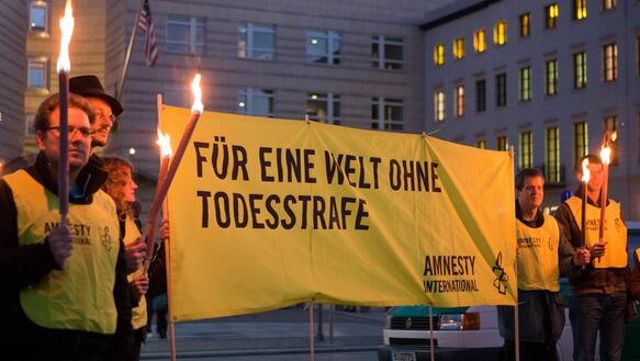 Gelb gekleidete Aktivisten mit Fackeln und einem Transparent gegen die Todesstarfe, dahinter ein Gebäude