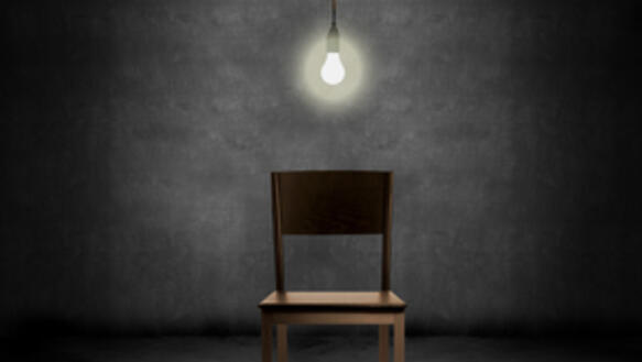 Ein Stuhl steht in einem leeren sonst dunklen Raum direkt unter einer hellen Glühbirne