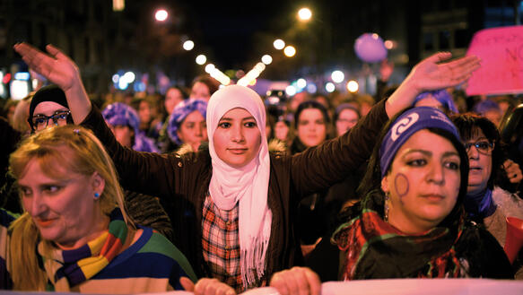 Viele Frauen laufen bei einer Demo, eine Frau mit weißem Kopftuch in der Mitte streckt die Arme zum Klatschen in die Luft