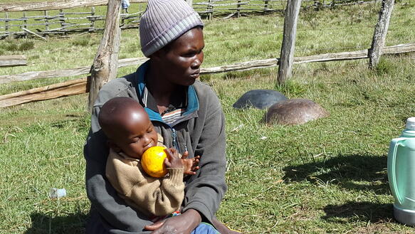 Eine Frau der kenianischen Sengwer sitzt mit einem Kleinkind auf dem Schoß in einer Wiese und blickt ernst in die Ferne.