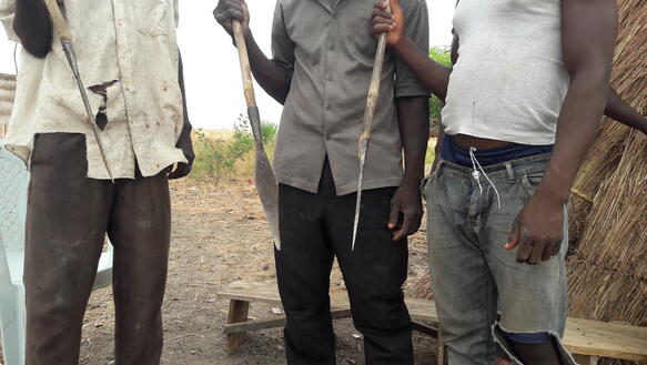 Drei Männer stehen vor einer Hütte und halten Speere in der Hand