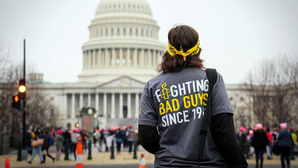 Eine Frau steht einem T-Shirt mit der Rückenaufschrift "Fighting Bad Guys" auf einer Straße vor dem weißen US-Senatsgebäude mit weißer Kuppel