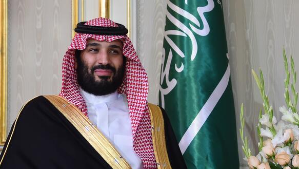 Porträt von Mohammed bin Salman vor der Saudi-Flagge und weißen Blumen