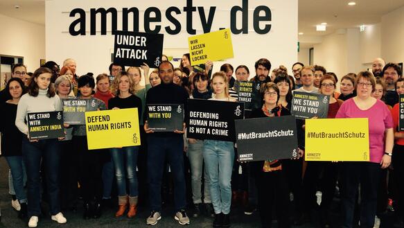 Eine Gruppe von Menschen hält schwarze und gelbe Plakate in die Höhe, im Hintergrund der Schriftzug amnesty.de.