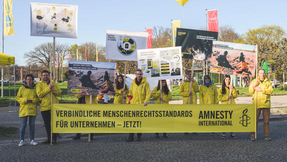Neun Personen in gelben Jacken stehen in einer Reihe und halten verschiedene Plakate und gelbe Luftballons hoch. Zusätzlich halten sie ein Banner mit der Aufschrift "Verbindliche Menschenrechtsstandards für Unternehmen - jetzt!"