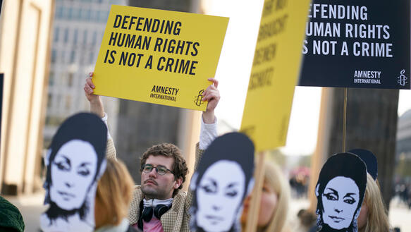 Eine Gruppe von Personen steht vor dem Brandenburger Tor und trägt Pappmasken mit dem Gesicht von Eren Keskin und hält Schilder und Banner in den Händen. Im Fokus ist ein Schild mit der Aufschrift „Defending human Rights is not a crime“