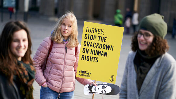 Drei junge Frauen stehen lächelnd im Kreis. Eine hält ein Pappschild mit der Aufschrift „Turkey: Stop the crackdown on human rights“