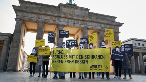 Eine Gruppe von Personen steht vor dem Brandenburger Tor und trägt Pappmasken mit dem Gesicht von Eren Keskin und hält Schilder und Banner in den Händen