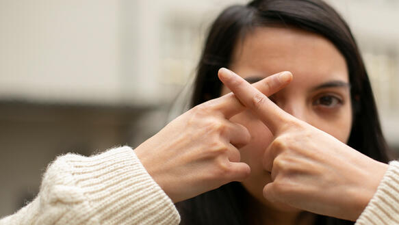 Eine junge Frau formt mit ihren Zeigefingern ein X vor ihrem rechten Auge und schaut direkt in die Kamera