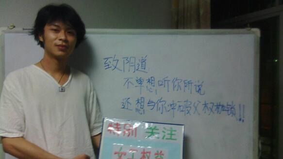 Ein junger Mann aus China steht in einem weissen T-Shirt vor einer Tafel mit chinesischen Zeichen und hält eine Botschaft auf Chinesisch in die Kamera