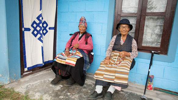 Zwei Frauen mit Hüten sitzen vor einem blauen Haus.