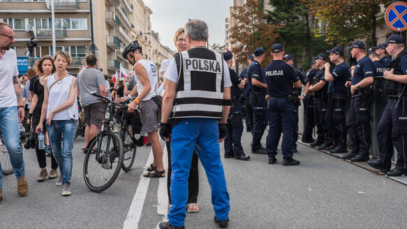 Links demonstrierende Menschen, rechts eine Reihe von Polizisten, dazwischen ein Mann mit einer Jacke, auf der "Polska" steht.