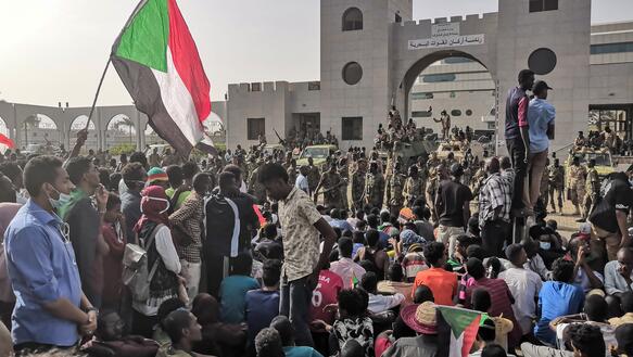 Eine Menschenmenge, zum Teil sitzend, eine hochgehaltene Fahne, dahinter Soldaten vor einem Tor