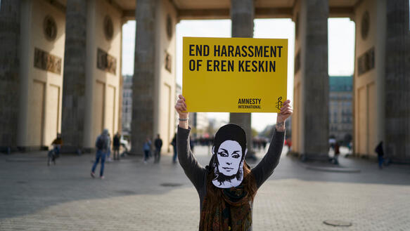Eine Frau mit Eren-Keskin-Maske steht vor dem Brandenburger Tor und hält ein Schild hoch mit der Aufschrift "End Harassen of Eren Keksen"