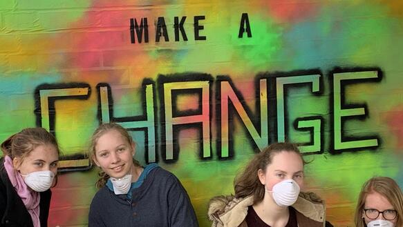Mehrere Jugendliche hocken mit Atemschutzmasken vor einem knallbunten Graffito mit dem Slogn "Make a change"