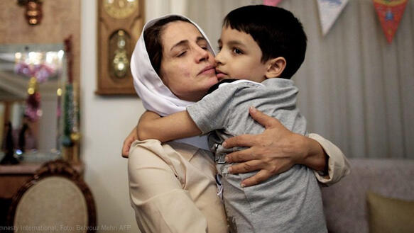 Eine Frau mit einem weissen Kopftuch  in einem Wohnzimmer drückt liebevoll ihren jungen Sohn an sich