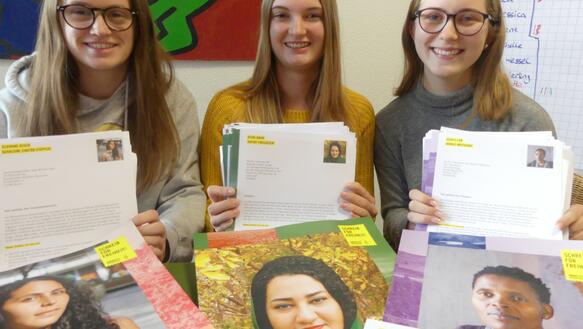 Drei Jugendliche halten Briefe in der Hand und vor ihnen liegen Porträtfotos verschiedener Frauen auf einem Tisch