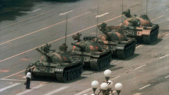 Vier Panzer fahren hintereinander auf einer breiten Straße auf einen Mann mit schwarzer Hose und weißem Hemd zu