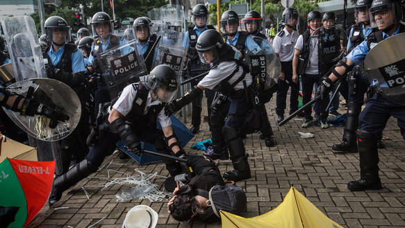 Ein Polizist in Schutzausrüstung schlägt auf eine Demonstrantin ein, die gekrümmt am Boden liegt, während mehr als zwölf Polizisten mit Helmen, Schilden und Schlagstöcken drumherum stehen