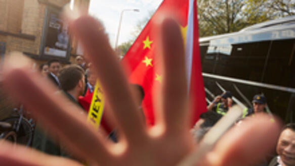 Eine Hand verdeckt teilweise die Kamera. Im Hintergrund befinden sich protestierende Menschen, welche die chinesische Flagge schwenken. 