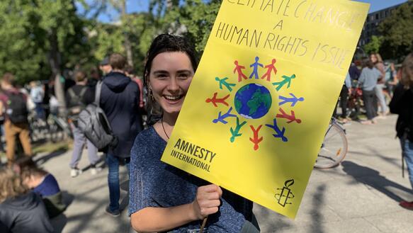 Eine junge Frau hält lächelnd ein Plakat in die Kamera, auf dem steht "climate change is a human right issue". Im Hintergrund steht eine Menschenmenge. 