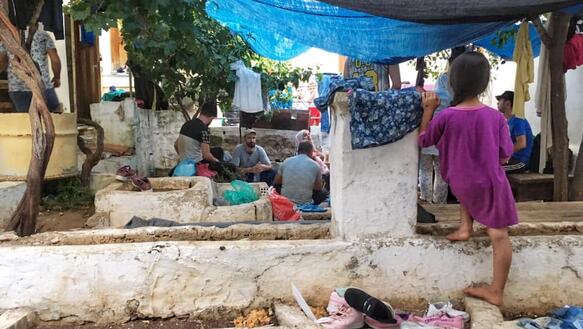 Erwachsene und Kinder befinden sich in einer provisorischen Unterkunft, dessen Dach aus Tüchern besteht. Auf dem Boden sind Kleidungsstücke verteilt.