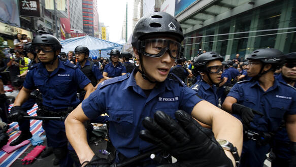 Eine Gruppe von rund zehn Polizisten mit Schlagstöcken, Helmen, Hemden und Schutzbrillen bahnt sich ihren Weg durch eine Straße, der vorderste Polizist steht ganz nah vor der Kamera