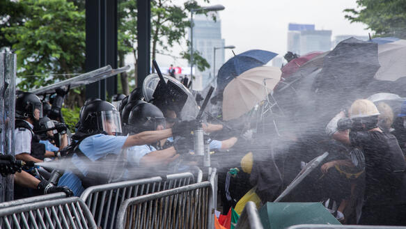 Eine Gruppe Polizisten in Schutzausrüstung sprüht über Absperrgitter hinweg Pfefferspray auf Demonstrierende, von denen sich einige mit Regenschirmen schützen