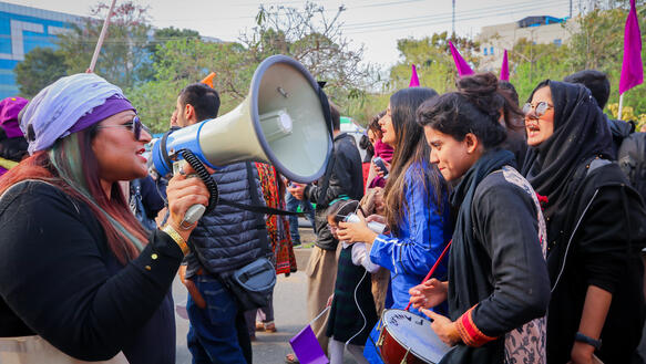 Eine Menschengruppe, die meisten von ihnen Frauen, zieht mit Fahnen und Trommeln durch eine Straße, die Frau im Vordergrund hält ein Megafon in der Hand