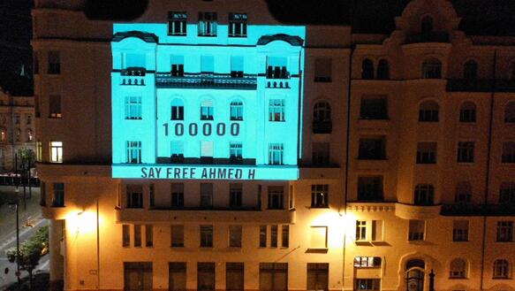 Auf einem Gebäude, welches wegen der Tageszeit beleuchtet ist, wird ein Bild projiziert, auf dem steht: "100.000 Menschen fordern: Lasst Ashmed H. frei"