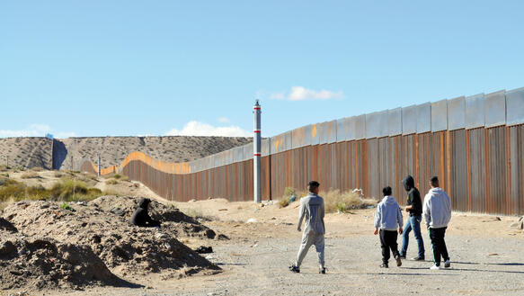 Eine hohe Mauer durchtrennt eine Landschaft, davor eine Gruppe von Jugendlichen.