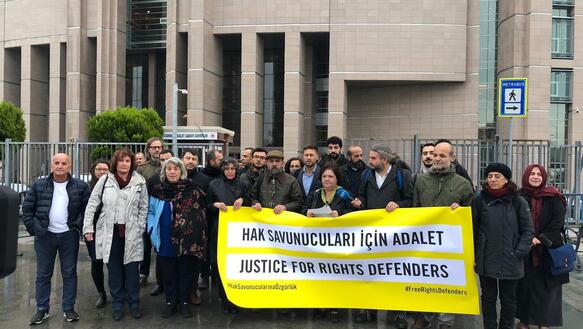Eine Gruppe von circa 25 Personen steht vor einem Gebäude, einige von ihnen halten ein Banner mit Forderungen in türkischer Sprache vor sich