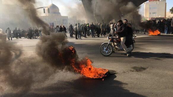 Eine Menge demonstrierender Menschen steht auf einer Straße im Iran. Die Menschen sind umgeben von Rauchwolken, die von mehreren Bränden ausgehen. Einige der Demonstrierenden sind auf Motorrädern unterwegs. 
