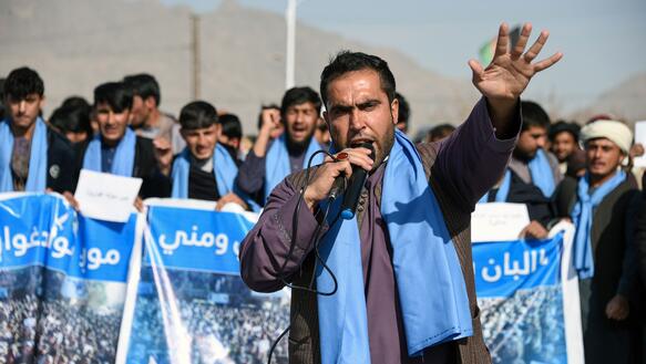 Mehrere Menschen mit blauen Plakaten protestieren, im Vordergrund ein Sprecher mit Mikrofon
