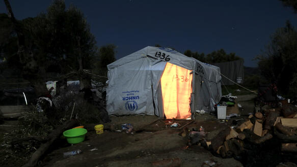In der Mitte des Bildes ist ein großes weißes UNHCR-Zelt zu sehen, aus dem es hell leuchtet, rings herum liegt Müll und verschiedene Gegenstände. Im Hintergrund sind Bäume zu sehen, es ist Nacht und dunkel.