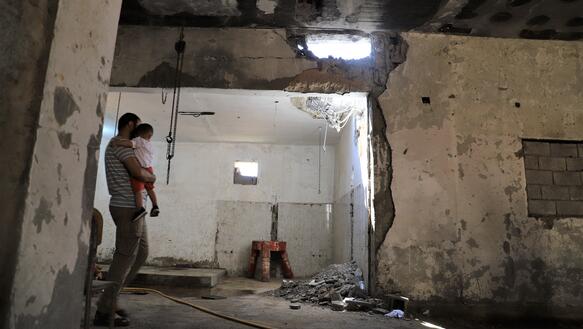 Ein Mann steht mit einem kleinen Kind auf dem Arm in einem zerstörten Haus. Die Wände sind schwer beschädigt, in der Decke ist ein Loch.