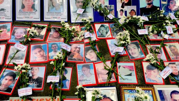 Fotos von getöteten Menschen liegen nebeneinander an einer Gedenkstätte, dazwischen Blumen