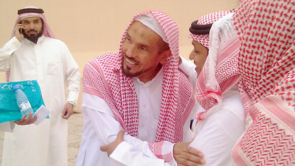 Vier Männer stehen vor einem einstufigem Gebäude. Sie tragen alle weiße saudi-arabischen Gewändern und rot-weißen Kopfbedeckungen. 