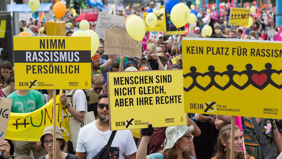 Eine Menschenmenge, aus der Luftballons und Schilder herausragen. Die vorderen Schilde sind schwarz-gelb. Auf ihnen steht: "Nimm Rassismus persönlich", "Menschen sind nicht gleich, aber ihre Rechte" und "Kein Platz für Rassismus"